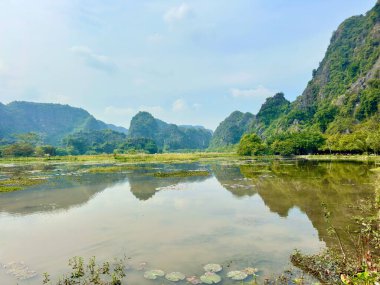 Mükemmel simetriyi yansıtan kireçtaşından çıkıntıları olan sulak bir vadinin panoramik görüntüsü. Nehir yüzeyi, gökyüzünü yansıtan dev bir ayna gibi davranır ve Tam Coc 'un kıyıları boyunca yeşil ağaçları yeşertir. Trang An Manzarası Şirketi' ndeki Bich Dong.