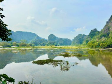 Yuvarlanan dağ sıralarının ortasındaki huzurlu sulak arazinin panoramik manzarası. Yeşil su sümbülleriyle benekli uçsuz bucaksız bir alan. Uzakta dalgalanan kireçtaşı dağları yüksek gökyüzünün altında bulanıklaşıyor. Tam Coc - Bich Dong esprisinde yakalandı