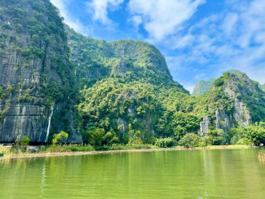 Tam Coc manzaralı yeşil nehri yansıtan görkemli kireçtaşı kayalıkları. Fotoğraf, kayalara yapışmış yosunlu ve ağaçlı karst jeolojik güzelliğini yakına çekiyor..
