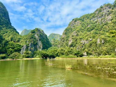 Ninh Binh 'deki Tam Coc Bich Dong turistik alanındaki huzurlu ve şiirsel manzaranın panoramik görüntüsü. Görüntü zümrüt nehri yakalıyor görkemli kireçtaşı dağlarının arasından geçiyor parlak mavi bir gökyüzünün altında beyaz bulutlarla..