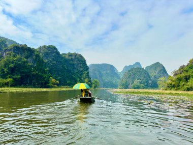 Bir kayıkçı, şiirsel ve manzaralı Ngo Dong Nehri 'ndeki huzurlu güzelliği keşfetmek için turistleri götürür. Fotoğraf, belirgin sarı ve yeşil şemsiyeli bir kayığın arkasından çekildi. Tam Coc - Bich Dong; Ninh Binh; Vietnam 10 Aralık 2025