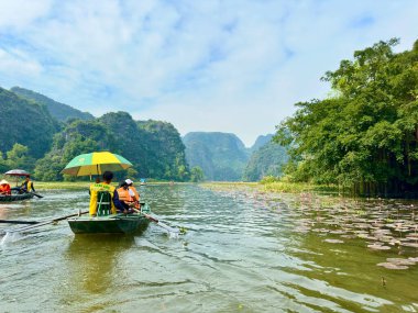 Turistler, tarihi Ngo Dong Nehri boyunca geleneksel el yapımı teknelerle güzel doğal manzaranın tadını çıkarıyorlar. Kayıkçının arkasından çekilen görüntü doğaya ve su yoluna ne kadar yakın olduğunu gösteriyor. Tam Coc - Bich Dong, Vietnam 10 Aralık 2025