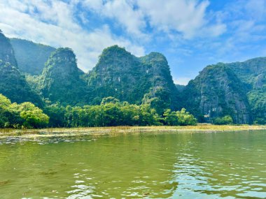 Ninh Binh topraklarındaki sessiz nehir kenarındaki dalgalı kireçtaşı dağlarının panoramik manzarası. Geniş açı, nehir boyunca uzanan sıradağların etkileyici bir doğal koridor oluşturduğunu gösteriyor. Tam Coc - Bich Dong