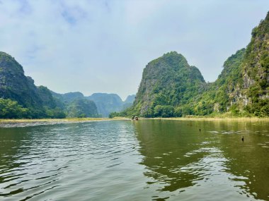 Görkemli kireçtaşı karst dağları Ninh Binh 'in manzaralı manzarasında huzurlu Ngo Dong nehrinin üzerinde yükseliyor. Görüntü, su bitkileriyle benekli sakin su yüzeyine yansıyan yuvarlanan dağların geniş açılı görüntüsünü yakalıyor.