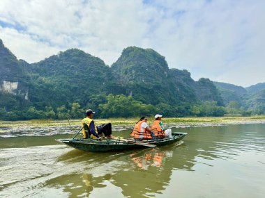 Bir bambu teknesinde oturan turistler Tam Coc Ninh Binh 'deki dağları seyrediyorlar. Turuncu can yelekli iki misafir mavi bir nehrin ortasında, yerli biri tarafından kürek çekilen bir teknede dinleniyorlar. Tam Coc - Bich Dong, Ninh Binh, Vietnam