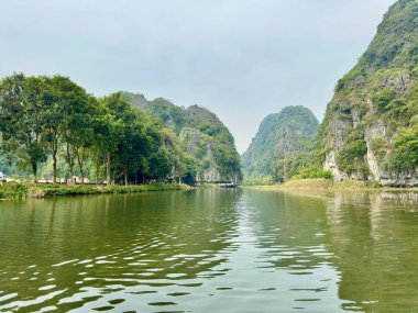 Tam Coc Ninh Binh 'de yemyeşil dağlara giden yolu açan huzurlu Ngo Dong Nehri. Fotoğraf, nehrin merkez manzarasından çekildi, kıyılardaki yeşil ağaçların suya yansıdığı, gözü uzak dağ sıralarına götürdüğü yerden. Tam Coc -