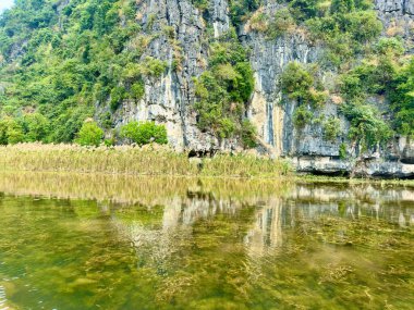 Dağ yansıması ve sazlıklar Ninh Binh 'in manzaralı bölgesindeki durgun gölde yansıyor. Fotoğraf, berrak mavi suyun mutlak dinginliğini yakalar. Kıyı boyunca uzanan her uçurumu ve sarı sazlığı net bir şekilde yansıtır. Tam Coc - Bich Dong
