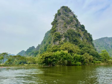 Tam Coc Ninh Binh sulak alanındaki yeşil ağaçlarla kaplı koni şeklinde kireç taşı dağı. Fotoğraf, yoğun tropikal bitki örtüsüyle kaplı nehrin kenarında duran ve suya yansıyan eşsiz şekilli dağa yakından bakıyor.