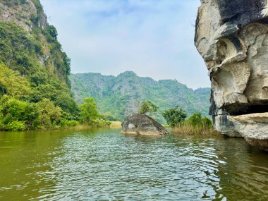 Tam Coc Ninh Binh miras alanındaki yeşil nehrin ortasında yalnız bir kaya. Fotoğraf nehir yatağından çıkan büyük bir kayayı gösteriyor. Yuvarlanan kireçtaşı dağları uzaklarda kayboluyor. Sessiz ve şiirsel bir his yaratıyor.