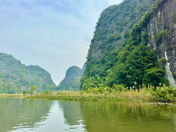 Ninh Binh bölgesindeki Tam Coc 'ta sarmal nehrin sakin güzelliği. Fotoğraf, sakin su yüzeyi, kıyı bitki örtüsü ve serin bir gökyüzünün altındaki yüksek gri kireçtaşı kayalıkları arasındaki zıtlığı tasvir ediyor. Tam Coc - Bich Dong