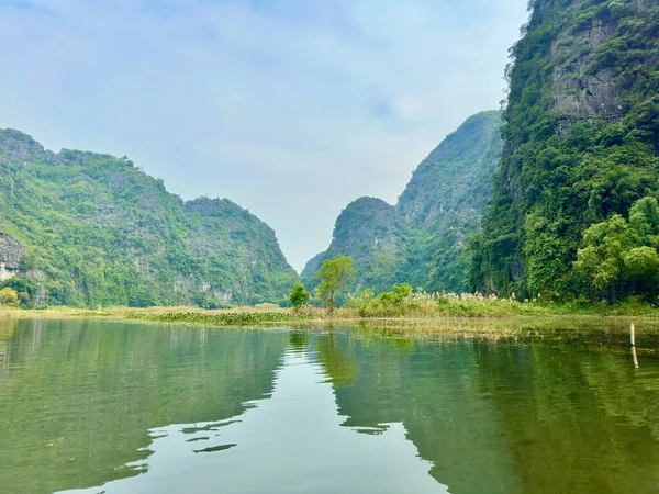 Sakin nehir yüzeyi, gökyüzünü ve uzak kireçtaşı sıradağlarını yansıtan bir ayna gibi hareket eder. Bu fotoğraf, su yüzeyinde yüzen su bitkilerinde bir açıklık ve boşluk hissi uyandırıyor. Tam Coc - Bich Dong, Ninh Binh