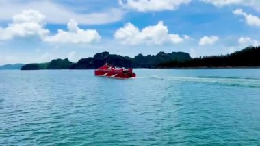 Canlı kırmızı yüksek hızlı feribot körfezin yemyeşil kireçtaşı dağlarında yol alıyor. Tekne turkuaz suya ve beyaz bulutlara karşı duruyor. Bai Tu Long Bay, Quang Ninh, Vietnam 16 Temmuz 2025. Video, hareketli hayatı yakalıyor.