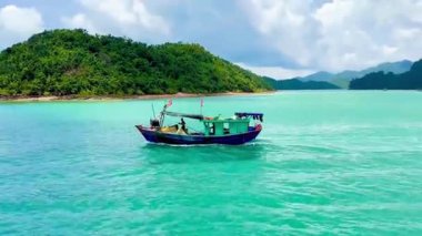 Koyun yeşil sularında süzülen yerel balıkçılardan oluşan ahşap bir balıkçı teknesi. Küçük tekne, çevreleyen yemyeşil adaların ihtişamıyla çelişiyor. Bai Tu Long Bay, Quang Ninh, Vietnam 16 Temmuz 2025. Görüntü yansıtır