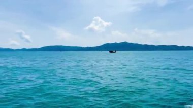 Vietnam 'ın turkuaz Bai Tu Long Körfezi bölgesinde yüzen küçük bir balıkçı teknesi. Görüntü, uçsuz bucaksız deniz ve gökyüzünün ortasındaki uzak sıradağlarla küçük, geleneksel bir ahşap kayığı gösteriyor. Bai Tu Long Körfezi, Quang Ninh, Vietnam