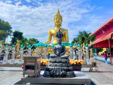 Pattaya 'daki Wat Phra Yai tapınağında altın Buda heykelinin önünde küçük bronz bir Buda heykeli. Arka planda büyük bir Buda heykeli ve para ağaçları olan bir kaideye bronz bir figür yakın. Büyük Buda Tapınağı Pattaya, Tayland