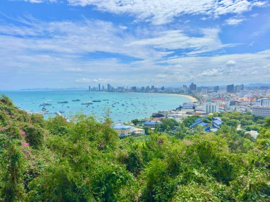 Pattaya sahil şehrinin şiirsel manzarası serin yeşil ağaç dalları ve yaprakları arasında belirdi. Yüksek açılı manzara kısmen yapraklarla kaplı. Mavi sahili ve uzaktaki yüksek binaları vurgulayan doğal bir çerçeve oluşturuyor..