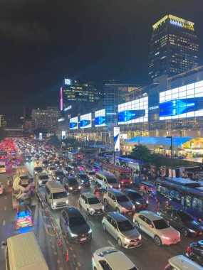 Bangkok 'taki CentralWorld alışveriş merkezinin önündeki hareketli sokak trafiğinin yüksek açılı görüntüsü. Devasa LED reklam ekranlarının parlak ışıkları altında sonsuz araba çizgileri uzanıyor. 29 Temmuz 2025 'te Bangkok, Tayland' da yakalandı..