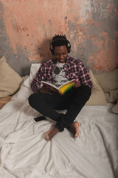 アフリカ系アメリカ人の若者がヘッドフォンをしてベッドの上に横たわって雑誌を読んでいます 高品質の写真 — ストック写真