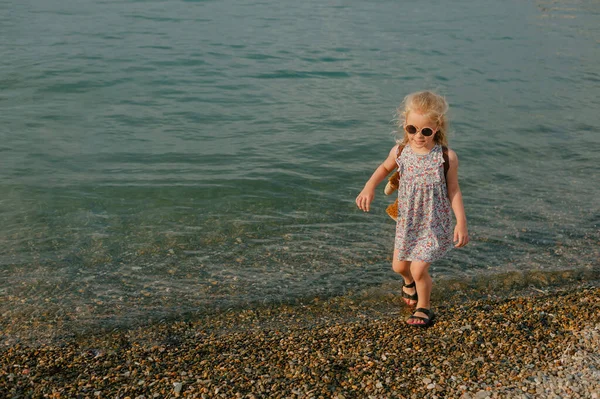 Glückliches kleines Mädchen in runden Gläsern spaziert im Sommer am Kiesstrand entlang — Stockfoto