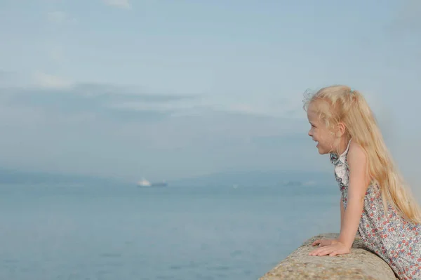 Klein meisje met lang wit haar in een jurk kijkt in de verte op de pier van de zee in de zomer — Stockfoto