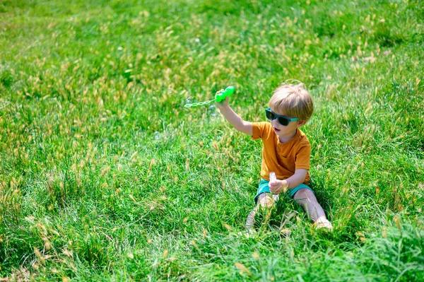 Petit garçon dans des lunettes de soleil en été est assis sur l'herbe et souffle des bulles — Photo