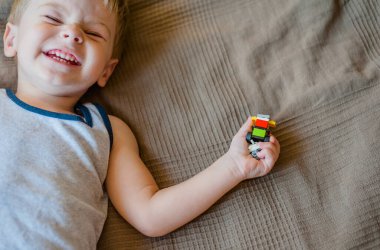 Küçük bir çocuk surat yapar, yatağa uzanır ve elinde bir Lego inşaatçısı tutar.