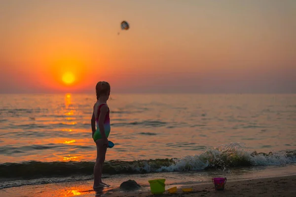 Silhouette d'une petite fille en maillot de bain au coucher du soleil debout dans la mer sur le fond de parasailing Images De Stock Libres De Droits