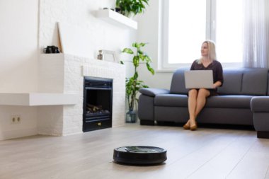 Kadın kanepede dinlenirken robot süpürgesi odayı temizliyor.