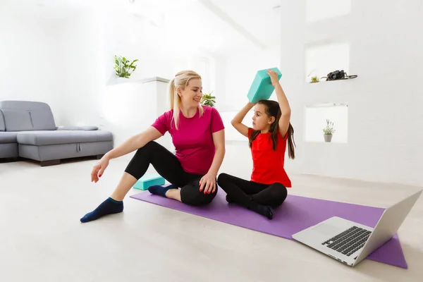 Mother and daughter practicing online yoga lesson at home at quarantine isolation period during coronavirus pandemic. Family doing sport together online from home. Healthy lifestyle
