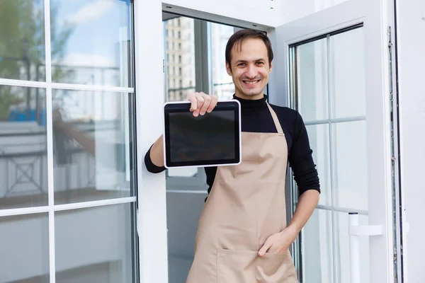 エプロンを着た顔の毛をした笑顔の若い男の肖像画とカフェでバーカウンターにもたれています テキストや画像のための空白のスレートボードのコピースペース バーテンダーの所有者のスタッフサービスと居心地の良いコーヒーショップ — ストック写真