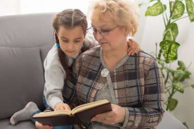 Mutlu küçük kız ve büyükanne kanepede oturup birlikte kitap okurlar.