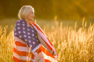 Çavdar tarlasında rüzgarda Amerikan bayrağı tutan güzel bir genç kız. Mavi gökyüzüne karşı yaz manzarası. Yatay yönelim.