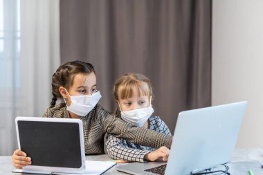 Mesafe öğrenme. Laptop 'ta Okul Kızları Maske Takıyor İnternette çalışıyor Evde masada oturuyor
