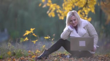 Autumn Park 'ta dizüstü bilgisayarı olan hoş bir kadın. Renkli yeşillik arka planlı güzel doğa sahnesi, sonbahar mevsiminde sarı ağaçlar ve yapraklar. Sonbahar açık hava yaşam tarzı. Mutlu gülen kadın güz yapraklarında