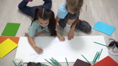 İki küçük kız kardeş evin zeminine uzanır ve renkli kalemlerle kağıt üzerine çizerler. Çocuklar okuldan sonra yaratıcı ödevler yaparlar..