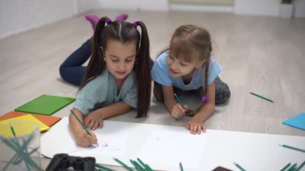 两个小女孩姐姐躺在房子的地板上 用彩色铅笔在纸上画画 孩子们放学后做创造性的家庭作业 — 图库视频影像