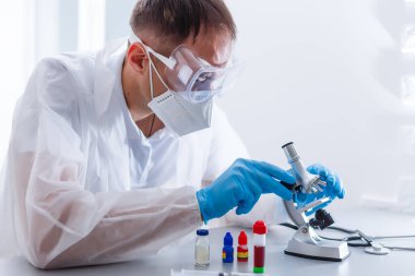 Arka planda dizüstü bilgisayarı olan bir erkek doktor ya da bilim adamı mikroskopla masaya bakıyor.