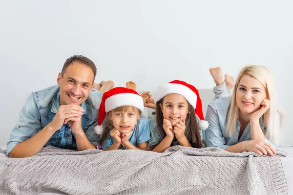 クリスマスサンタの帽子の家族はベッドの上に横たわっています 寝室で楽しんでる 家でリラックスしてる 冬の休日クリスマスと新年のコンセプト — ストック写真