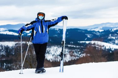 Kışın Coronavirus. Yüz maskesiyle kayak yapmak. Pandemia 'da kış tatili. Yüz maskeli beyaz bir gencin portresi. Kış yeni bir gerçeklikle geliyor..
