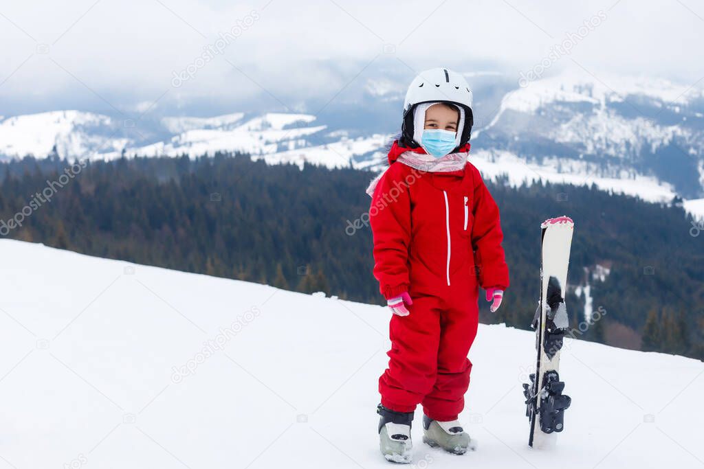 Portrait of a little girl skier in medical mask during COVID-19 coronavirus on a snowy mountain at a ski resort