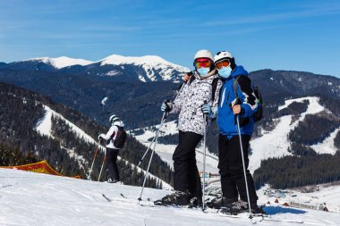 Skier wearing a medical mask during COVID-19 coronavirus in ski resort. Man and woman together in winter nature. Romantic scenery. Sport photo clipart