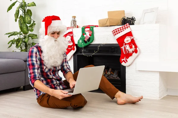 サンタクロースとノートパソコンはクリスマスのために装飾された部屋のテーブルに座って — ストック写真