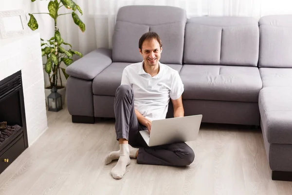 Young attractive smiling guy is browsing at his laptop, sitting at home on the cozy beige sofa at home, wearing casual outfit