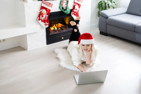 流行の冬のファッションでかなり若い女性は カメラを見ながら フロントクリスマスの装飾でノートパソコンガジェットを保持床に座って — ストック写真