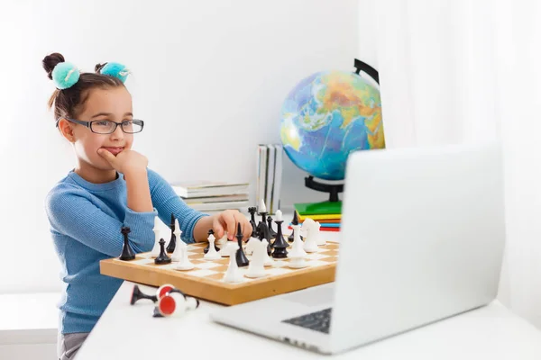 Portrait Caucasian Little cirly girl playing chess at the table in interior with chessboard. little girl learns chess on a laptop online, distance learning