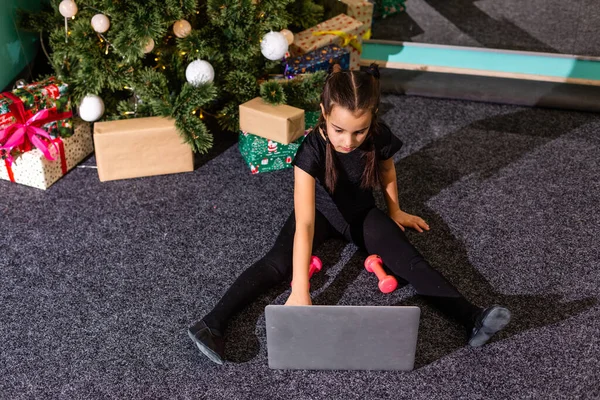 Little girl practicing gymnastics at home, watching online video on laptop. Social distance, online education concept. Healthy lifestyle, coronavirus, stay home. Kids sport home quarantine.