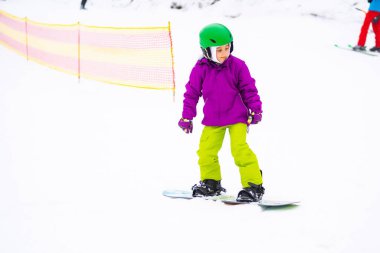 Snowboard Kış Sporu. Küçük kız snowboard öğreniyor, kalın kışlık giysiler giyiyor. Kış arkaplanı.