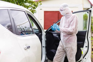 Kişisel koruyucu giysi, eldiven, cerrahi maske ve yüz maskesi takan bir adam, bir arabada oturan başka bir adam üzerinde kovid-19 koronavirüsü test ediyor..