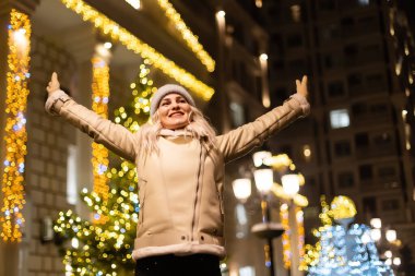 Açık havada, elinde maytap tutan, sokakta poz veren mutlu, genç bir kızın fotoğrafı. Arka planda şenlikli Noel panayırı. Şık bir kışlık manto, örgü şapka, fular....