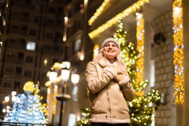 Açık havada, elinde maytap tutan, sokakta poz veren mutlu, genç bir kızın fotoğrafı. Arka planda şenlikli Noel panayırı. Şık bir kışlık manto, örgü şapka, fular....